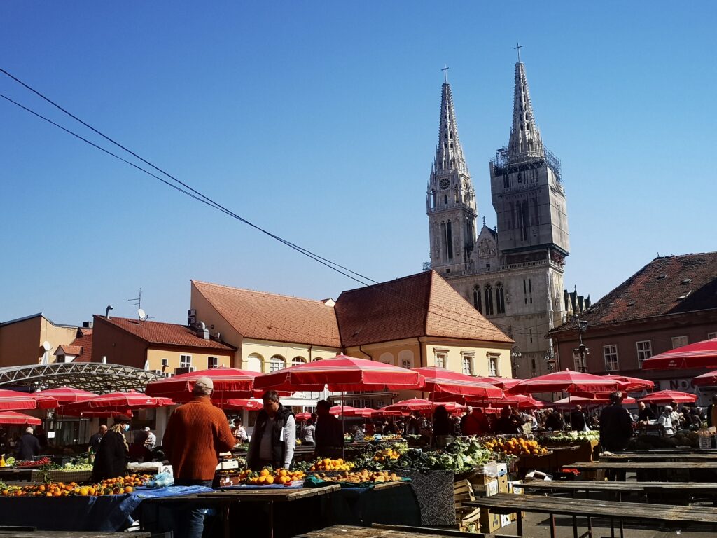 A view of the city landmark Zagreb Cathedral from inside the Dolac Market. The umbrellas are red and fresh fruit is being sold from their stalls.