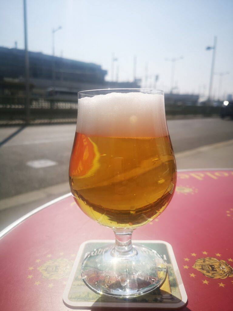 European beer served in a rounded glass, on a red table outside the bar, with a view of the station in the background.