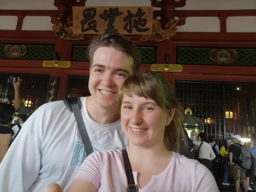 Myself (28F) and Partner (28M) stood inside the Sensoji Temple, with a wooden sign in Japanese above our heads and the inner temple behind us, protected by glass and metal fencing. One of the Top Tokyo Attractions.