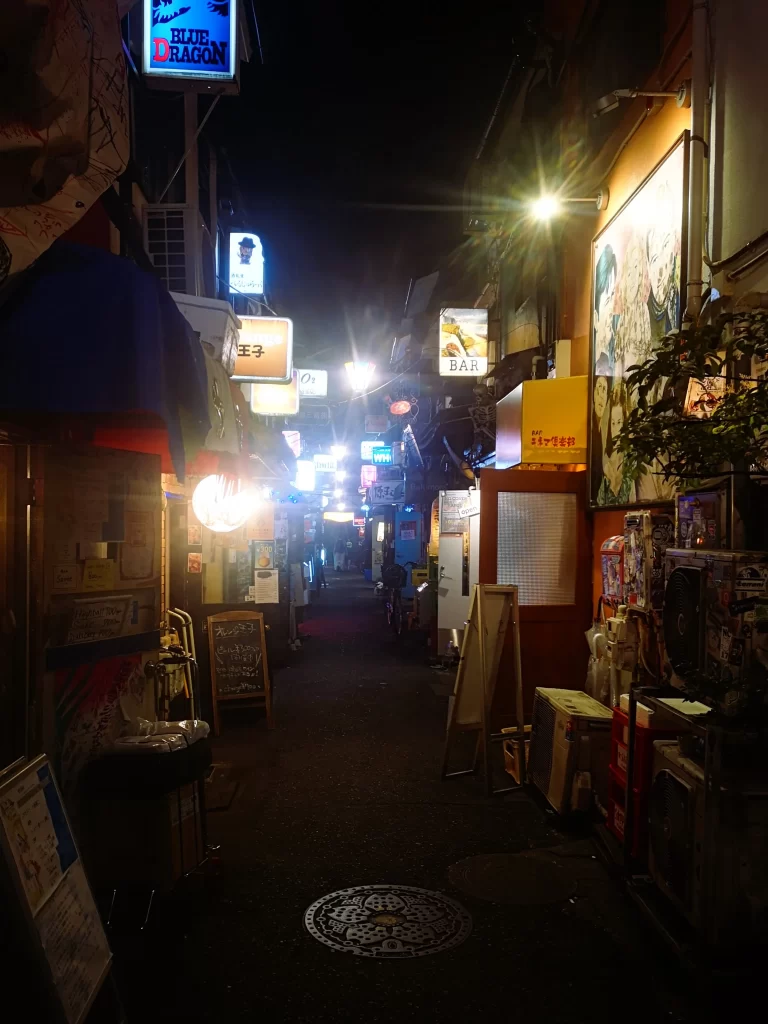 A long, very narrow street, filled with signs and lights and bar signs/boards outside the entrances. Several doors are open and there is not much space.