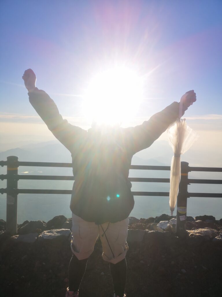 The sun is making my head invisible, at the top with celebratory arms in the air, celebrating climbing Mount Fuji